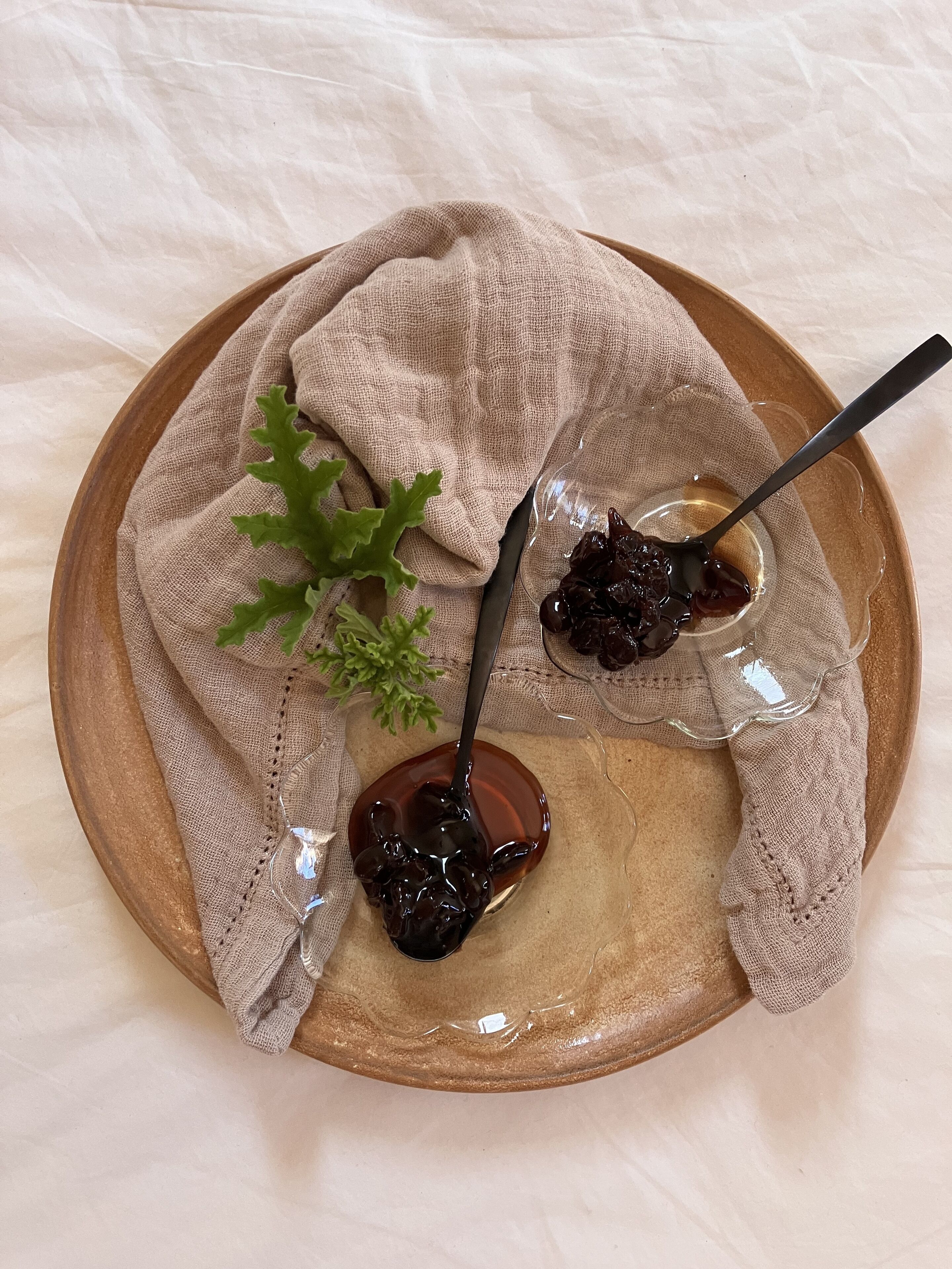 A wooden plate with two glasses of dark liquid, possibly a sauce or condiment, accompanied by a spoon and a piece of cloth.