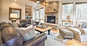A spacious living room with a stone fireplace, leather sofas, and large windows offering a forest view. - Luxury End Unit Townhome - Easy Access to Slopes - Views/Huge Deck (Durango)