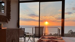 A bedroom with a large bed, a wooden dresser, a small table, and a chair. The room has large windows with a view of the sunset over the ocean. - Villa Asteria @ Mangata (Quepos)