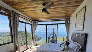 A bedroom with a bed, a ceiling fan, a nightstand, and a view of the ocean. - Villa Asteria @ Mangata (Quepos)