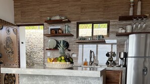 A modern kitchen with a central island, wooden stools, a stainless steel refrigerator, and a stone wall. - Villa Asteria @ Mangata (Quepos)