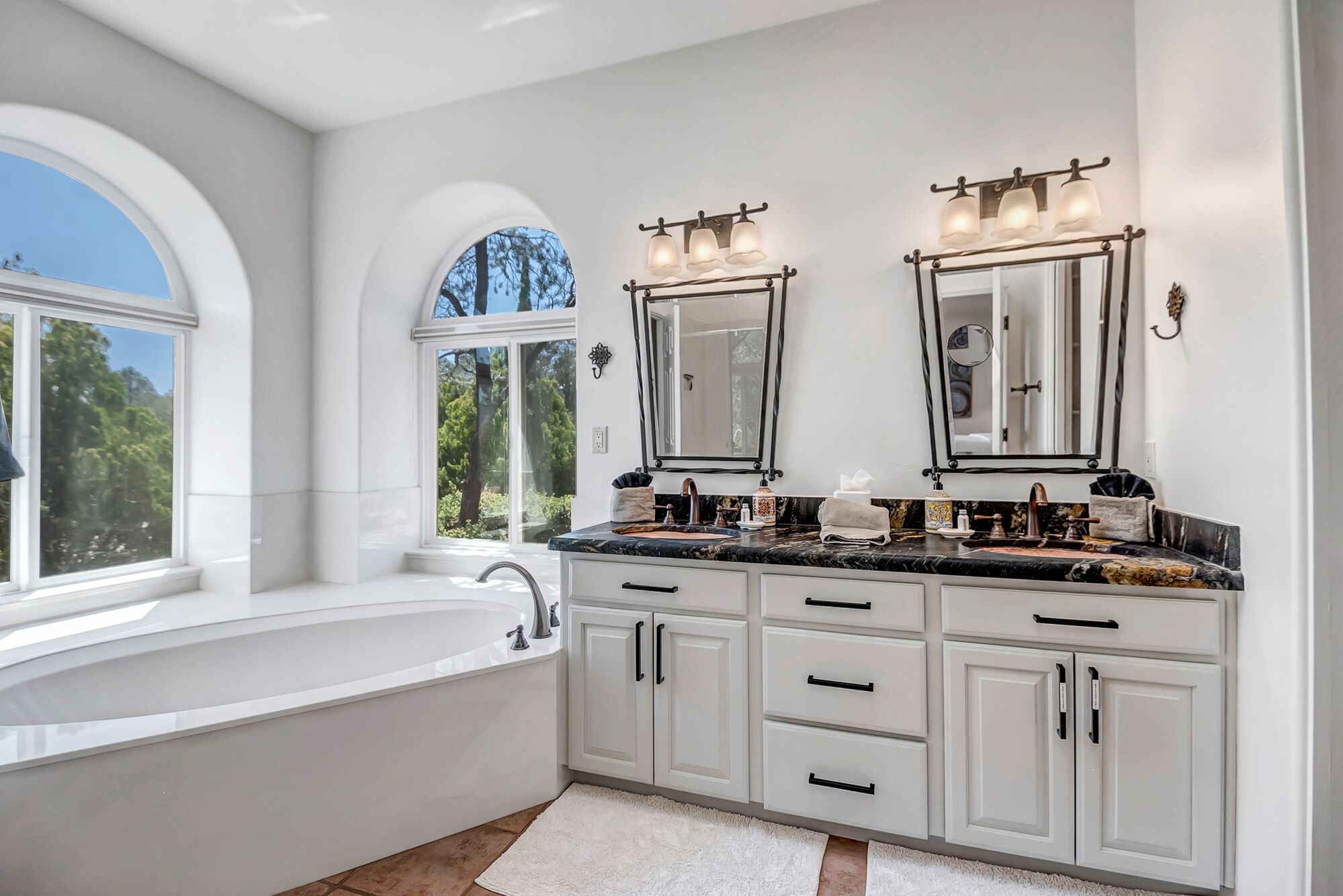 A spacious bathroom with a large white bathtub, double vanity with mirrors, and a window providing a view of trees.