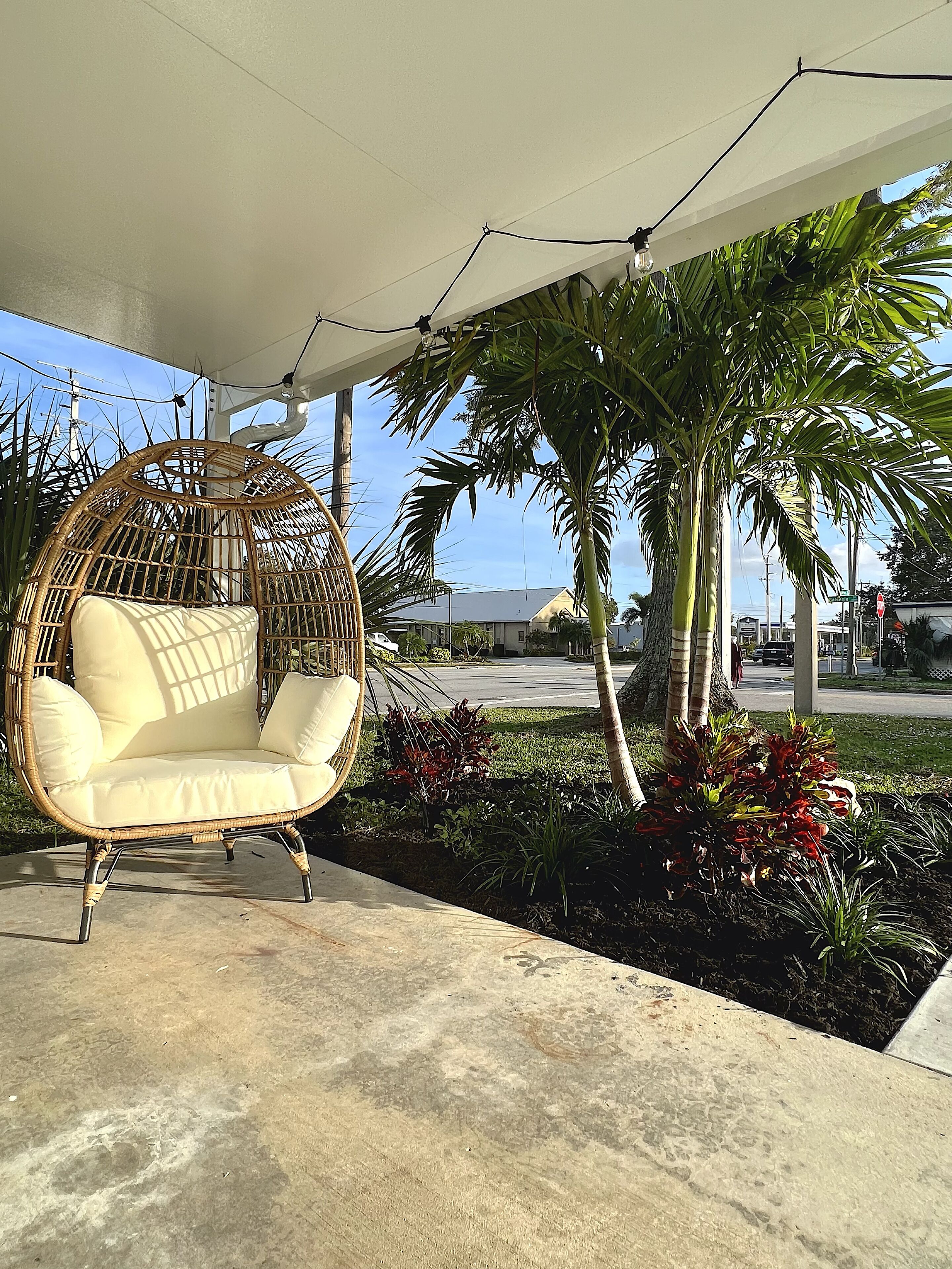 A wicker chair with cushions under a covered area, surrounded by tropical plants and palm trees.