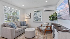 A modern living room with a grey sofa, ottoman, and armchair, a large flat-screen TV, and a view of the outdoors through large windows.