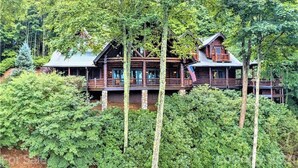 A log cabin with multiple decks and a porch, surrounded by dense forest. - 4BR / 4 1/2 BA,  Covered deck with Hot-tub overlooking the mountains (Maggie Valley)