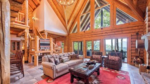 A spacious log cabin with a high ceiling, a fireplace, a cozy seating area, and large windows offering a view of the surrounding forest. - 4BR / 4 1/2 BA,  Covered deck with Hot-tub overlooking the mountains (Maggie Valley)