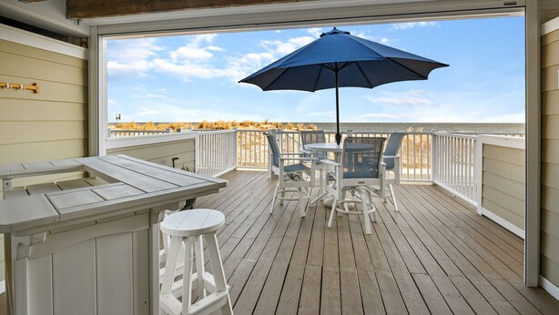 A wooden deck with a white railing, a table with chairs, and a blue umbrella. - Beach Walk II 6 by Seaside Vacations & Sales (Ocean City)