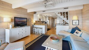 A modern living room with a TV, white cabinets, a sofa, and a staircase leading to a kitchen. - Beach Walk II 6 by Seaside Vacations & Sales (Ocean City)