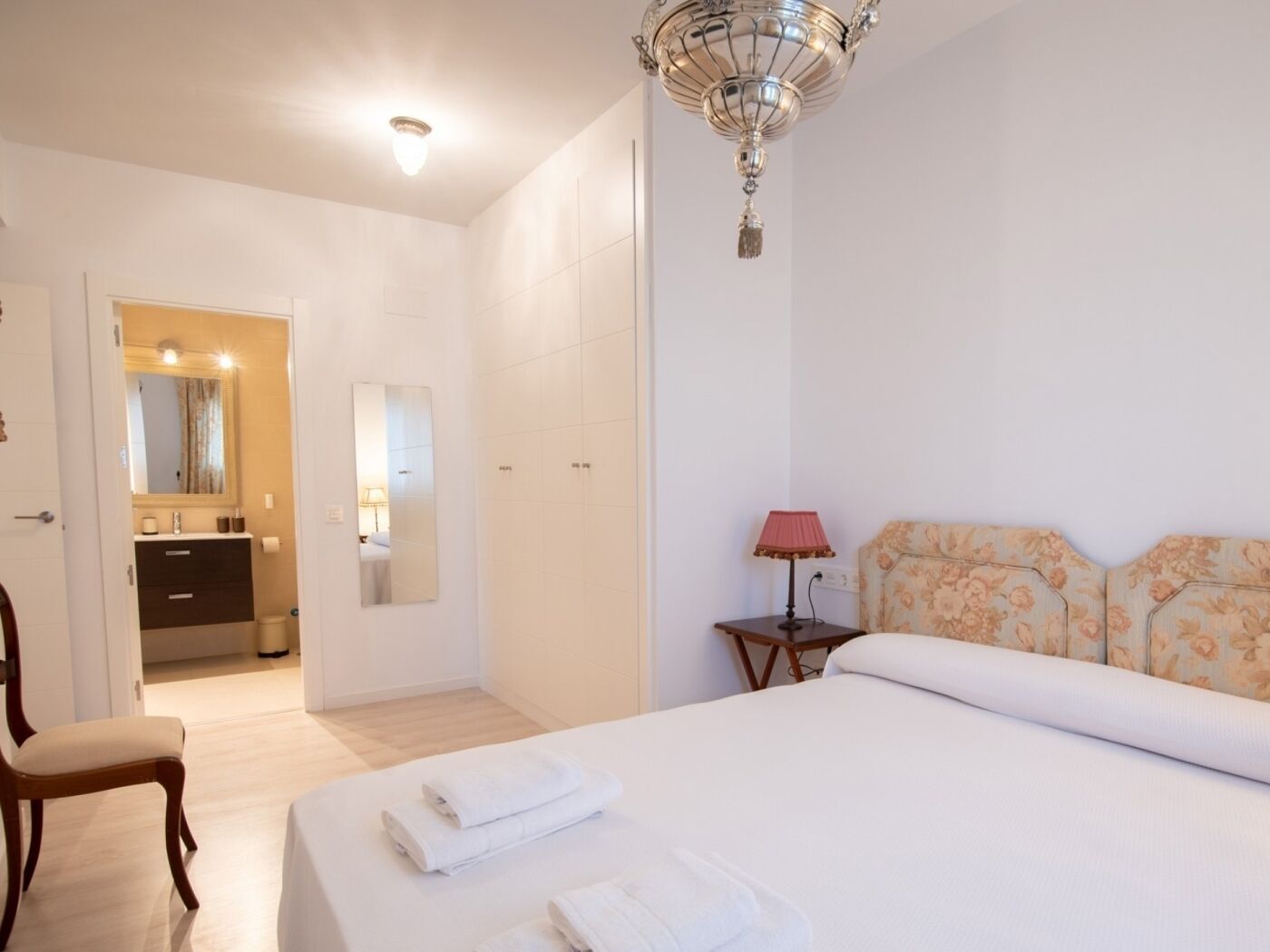 A hotel room with a bed, a chair, a bedside table with a lamp, a mirror, and a bathroom visible through an open door.