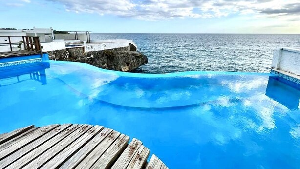 A wooden deck leading to a blue infinity pool with a view of the ocean and a cloudy sky. - ON THE CARIBBEAN SEA - CRYSTAL APARTMENT (Boca Chica)