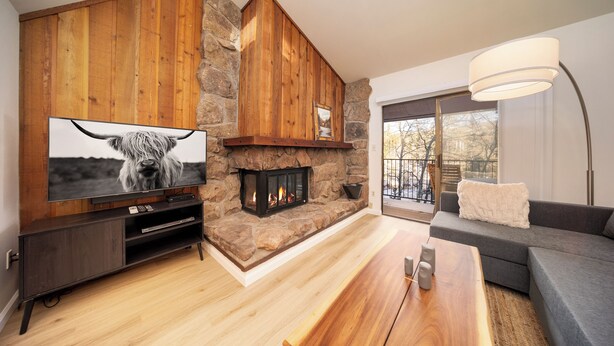A modern living room with a stone fireplace, wooden walls, a large flat-screen TV, a dark wooden entertainment unit, a grey sofa, and a large window with a view of trees. - Beaver Village Unit #0332 Remodeled 1 Bedroom vaulted ceiling & private deck (Winter Park)