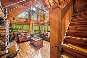 Une cabane spacieuse dotée d’une chambre à coucher en mezzanine, d’un salon confortable avec une cheminée et de grandes fenêtres offrant une vue sur la forêt environnante.