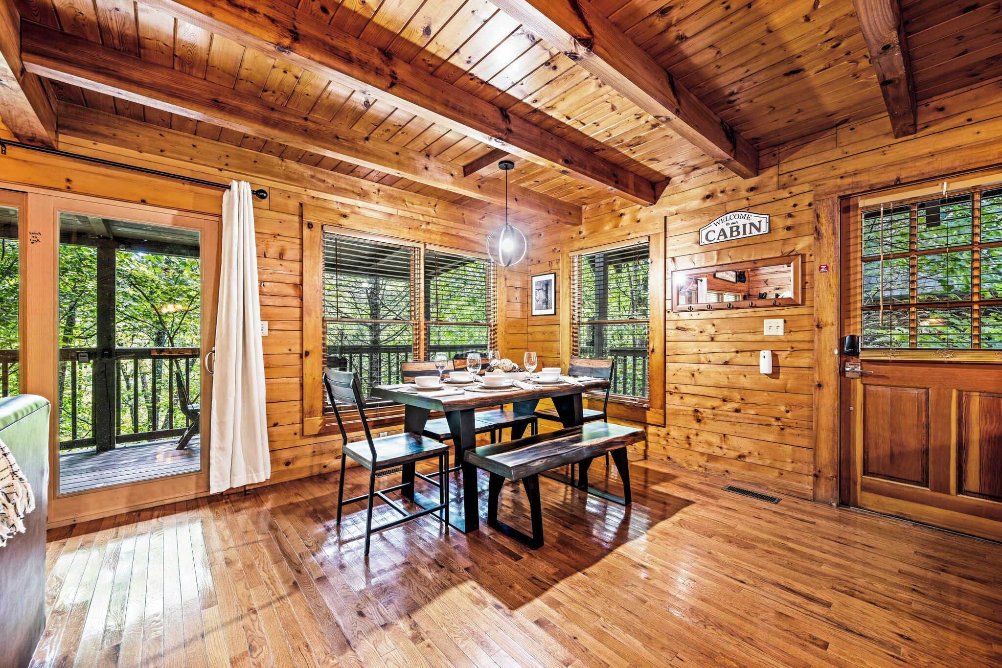 Een houten chalet van binnen, met een eettafel voor vier personen, grote ramen en een bordje met de tekst 'Welcome Cabin'.