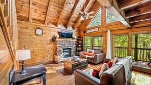 A cozy log cabin interior with a stone fireplace, wooden ceiling beams, a sofa, and a large window overlooking greenery. - PROMO 10% WEEKLY DISCOUNT September Special,Hot tub Pool table, Arcades WIFI-Pet (Sevierville)