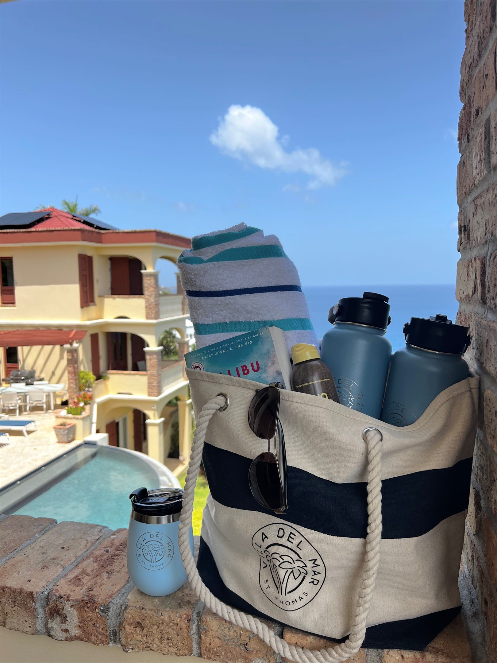 A beachside view with a pool, a house, and a bag with water bottles and a towel.