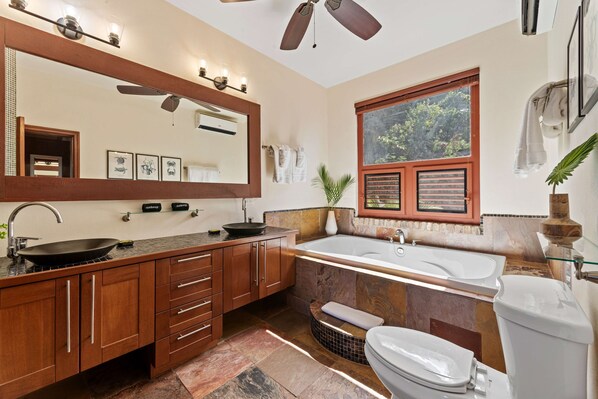 Un baño moderno con un tocador grande, una bañera independiente, un inodoro y una ventana con vista a follaje.
