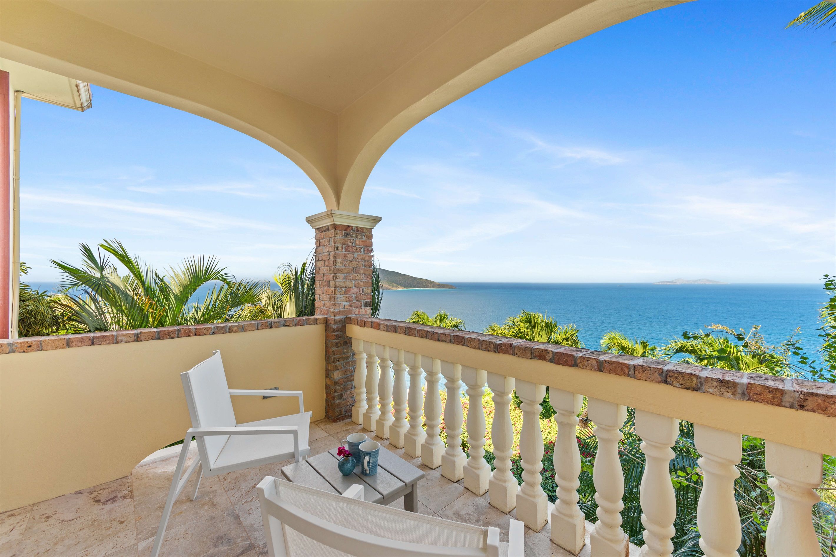 Un balcon avec vue sur la mer, des chaises blanches et une table agrémentée d’un vase et de tasses.