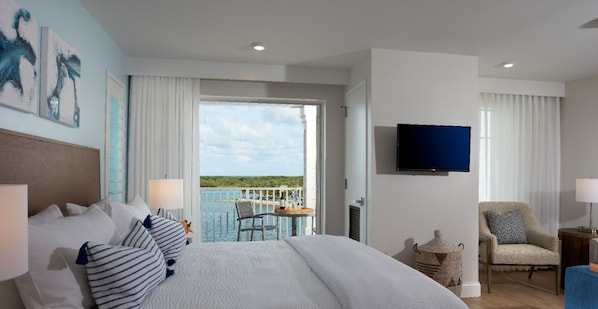 Habitación de hotel con una cama grande, televisión, balcón y vistas al mar, además de una silla.
