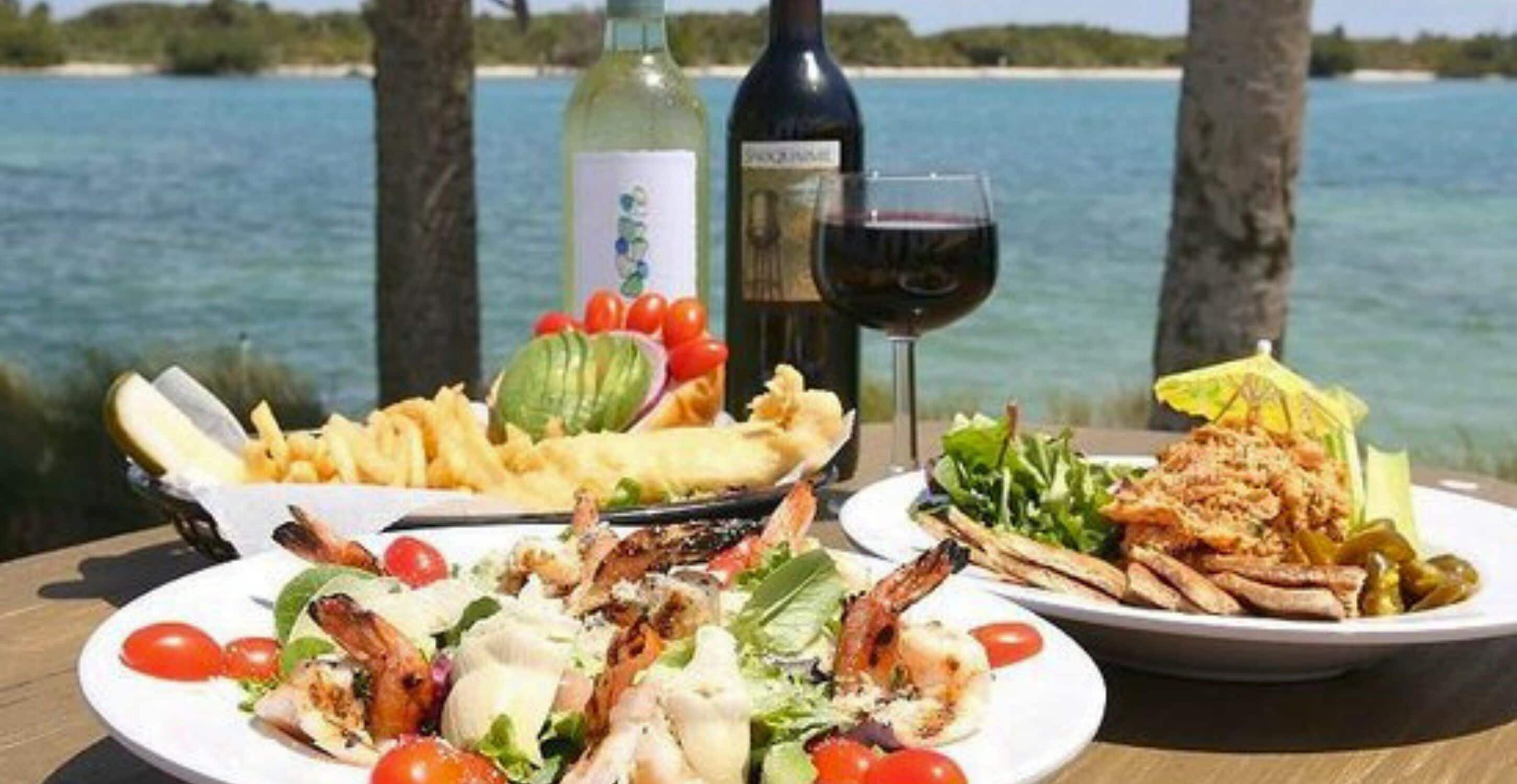 A table with seafood dishes, a bottle of wine, and a glass of red wine, set against a coastal backdrop.