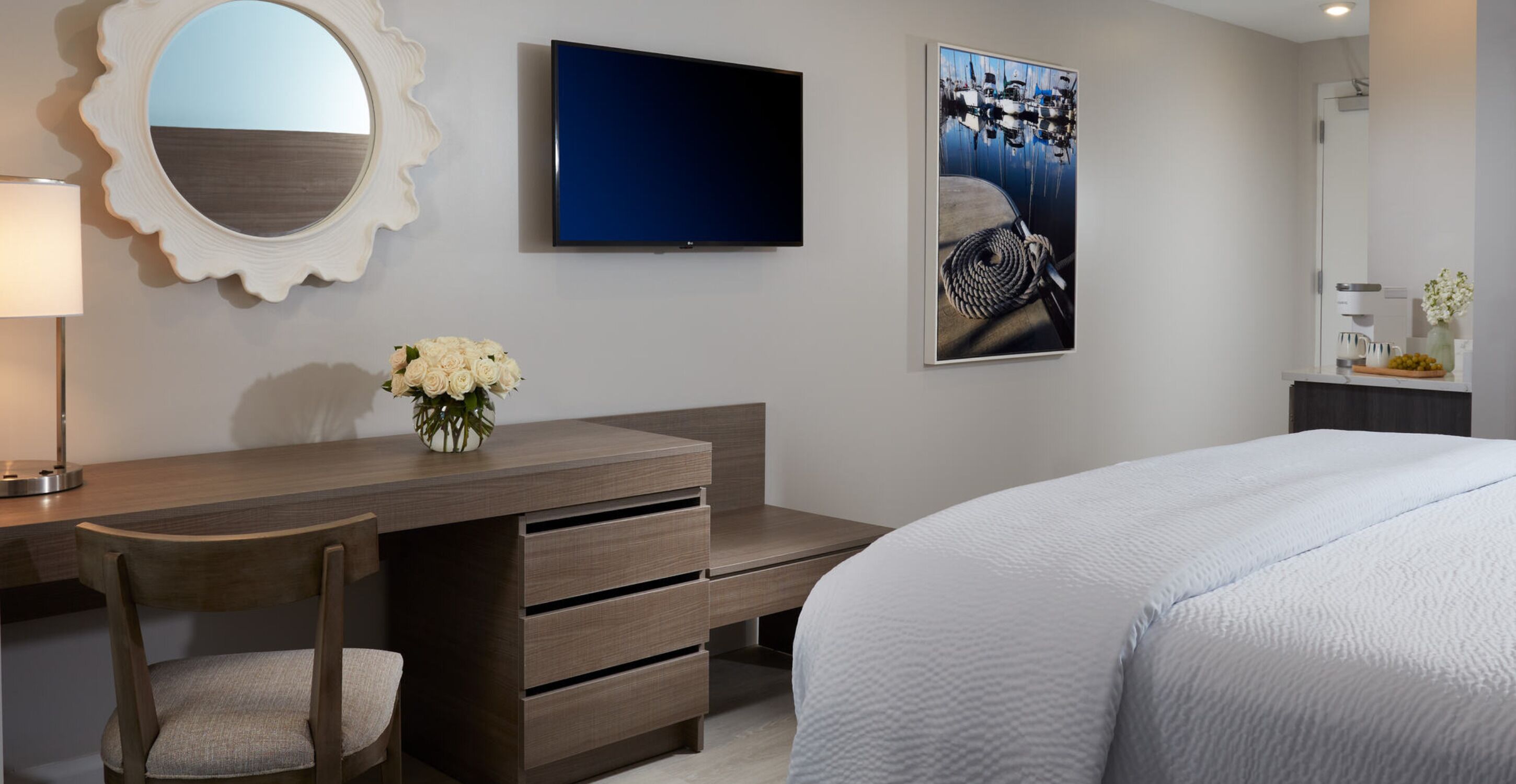 A modern hotel room with a bed, a desk with a chair, a TV, a mirror, and a framed picture on the wall.