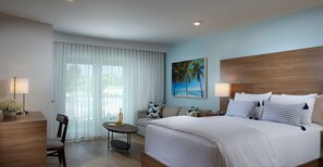 A hotel room with a large bed, a desk with a lamp, a chair, a small table with a bottle and glasses, and a painting of palm trees.