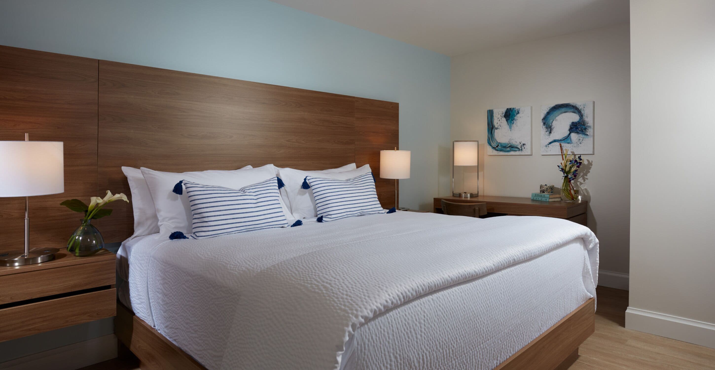Una cama bien hecha con almohadas a rayas blancas y azules, un cabecero de madera y una mesita de noche con una lámpara y un florero.