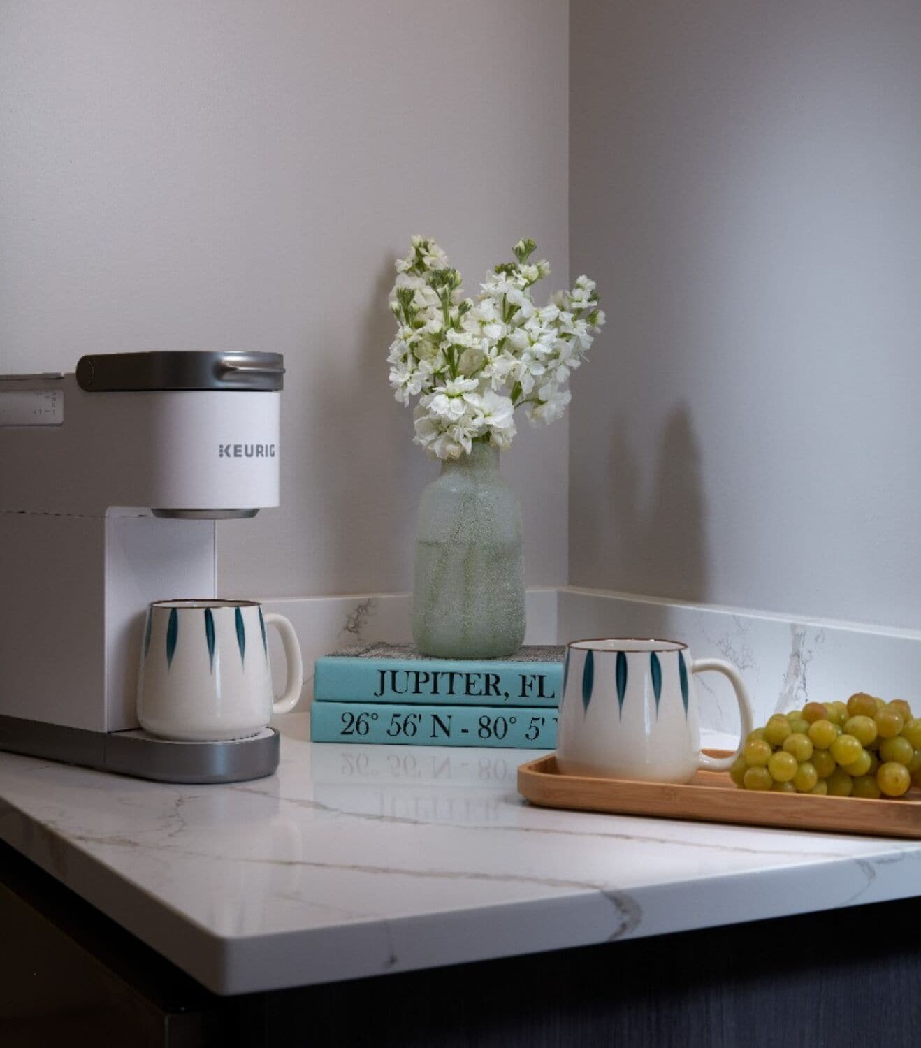 A kitchen counter with a Keurig coffee maker, a vase with white flowers, a mug, a book titled 'JUPITER, FL', and a bowl of grapes.