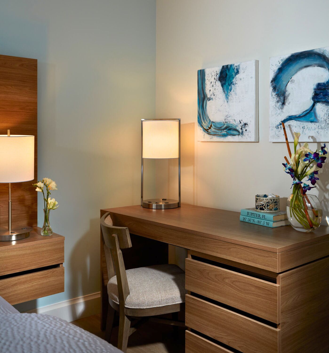 Une chambre d’hôtel avec un bureau en bois, une chaise, une lampe et deux tableaux abstraits accrochés au mur.