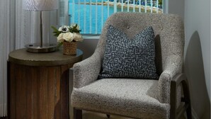 Una habitación con vista al mar, una silla con cojín estampado, una lámpara y una planta pequeña sobre una mesita de madera.