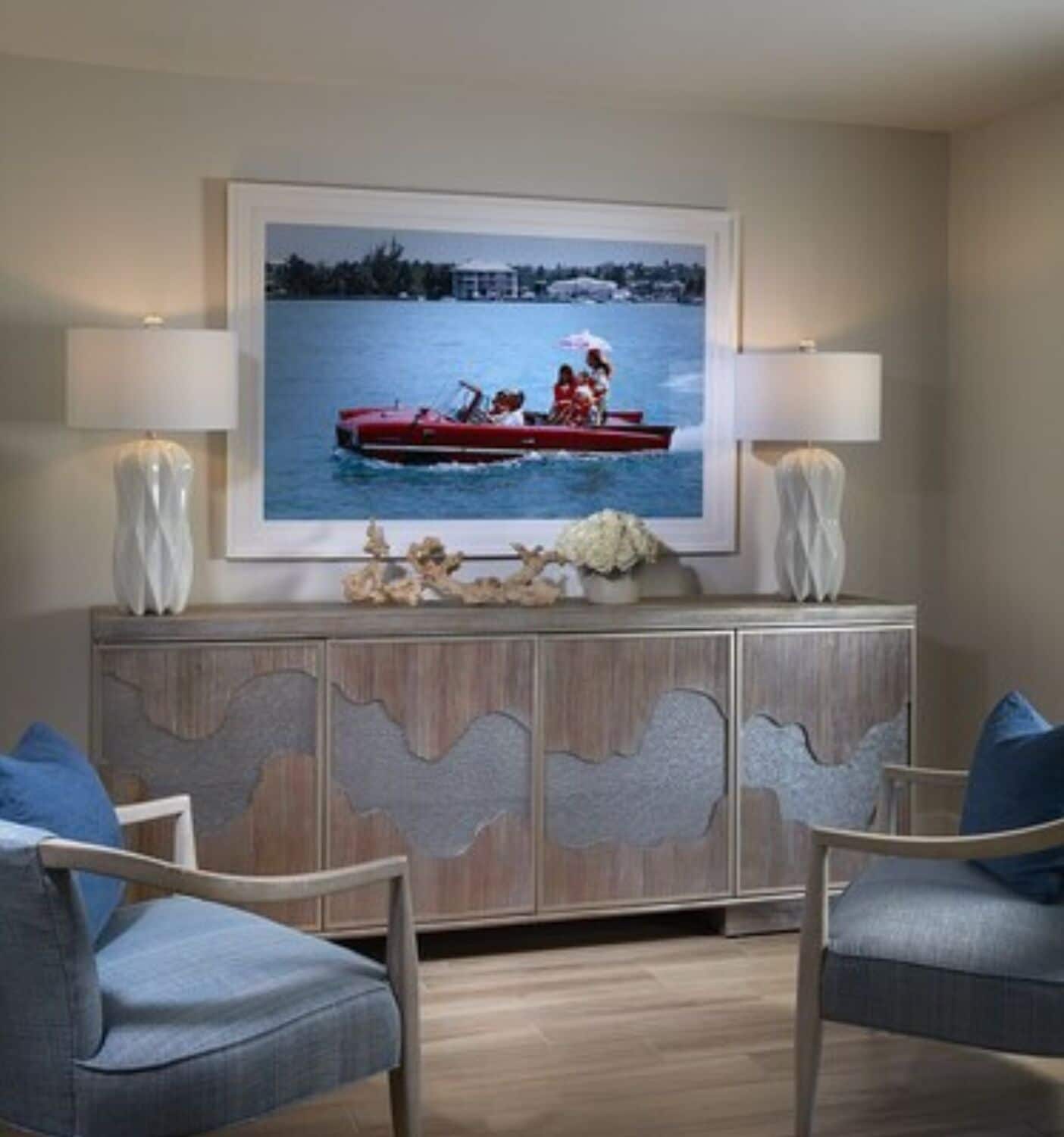 Uma sala de estar com um quadro grande de um barco vermelho na água, um armário de madeira com detalhes entalhados e duas poltronas azuis.