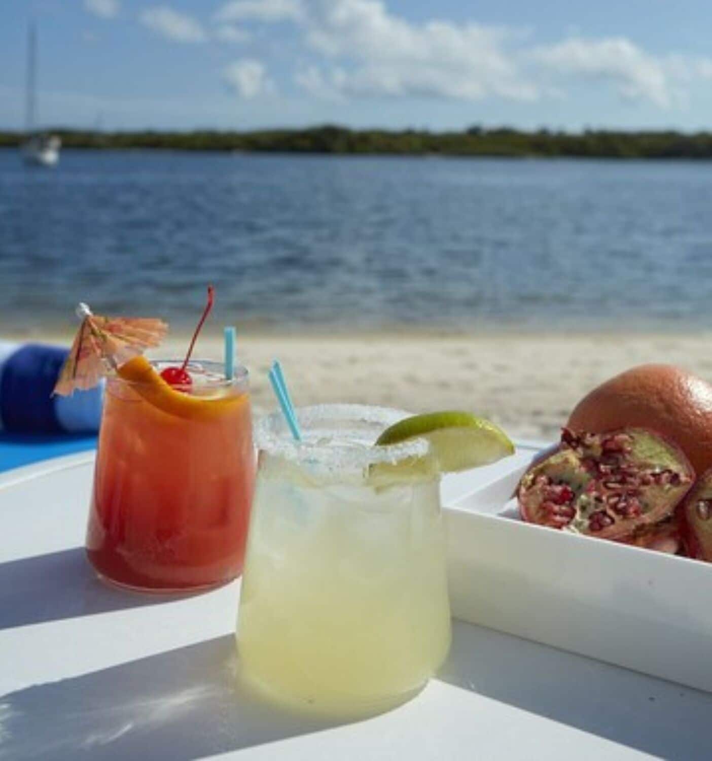 Una mesa con dos bebidas y un sándwich en una playa, con vista al agua.