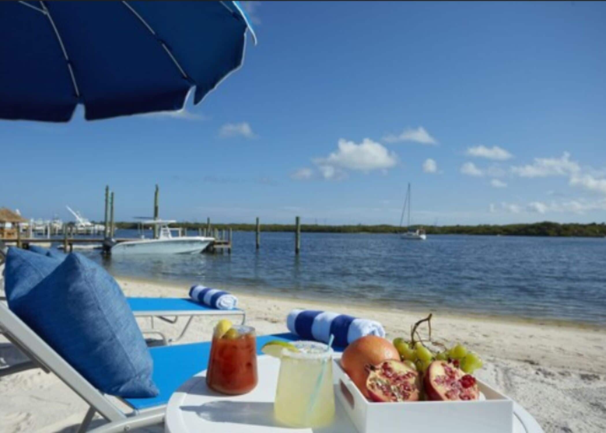 A beachside view with a table set for a meal, including fruits and drinks, under a blue umbrella.