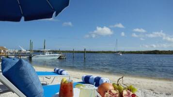 A beachside view with a table set for a meal, including fruits and drinks, under a blue umbrella.