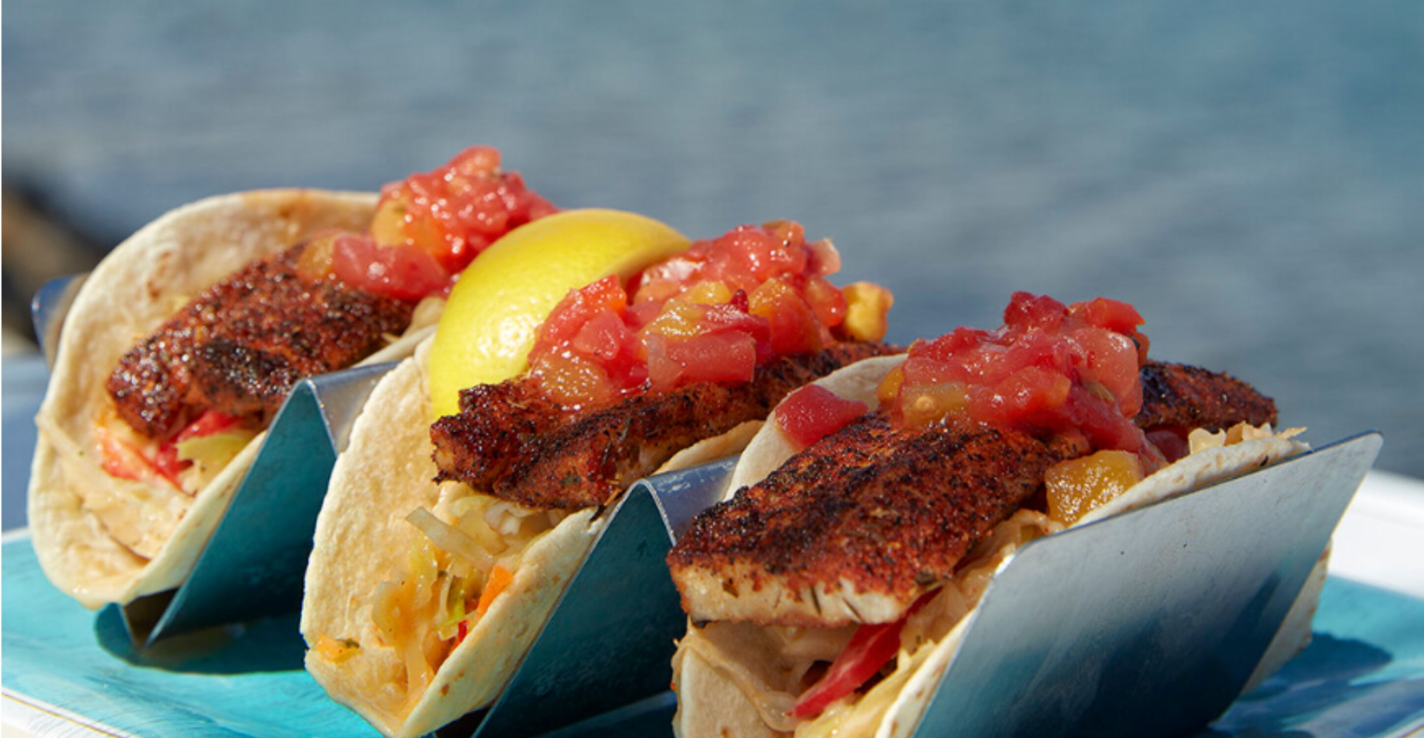 Three tacos with fish, tomatoes, and a lemon wedge.