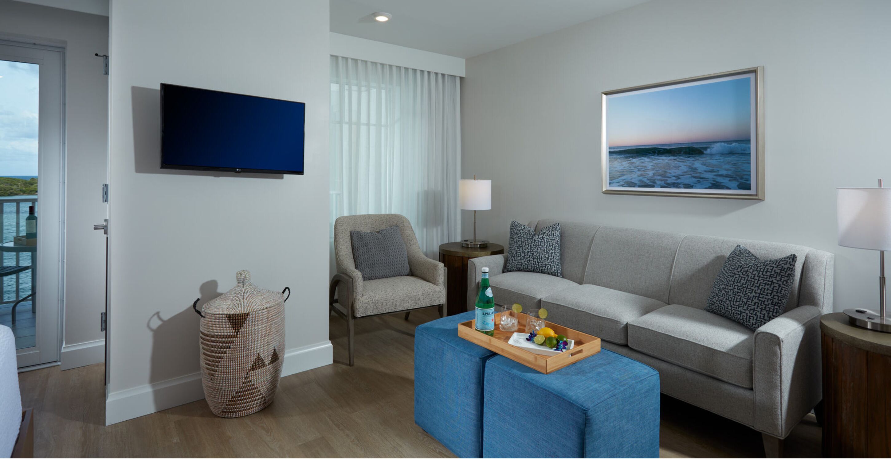 A modern living room with a grey sofa, a blue ottoman, a wicker basket, a television, and a framed picture of a beach.