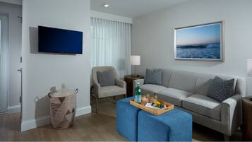 A modern living room with a grey sofa, a blue ottoman, a wicker basket, a television, and a framed picture of a beach.