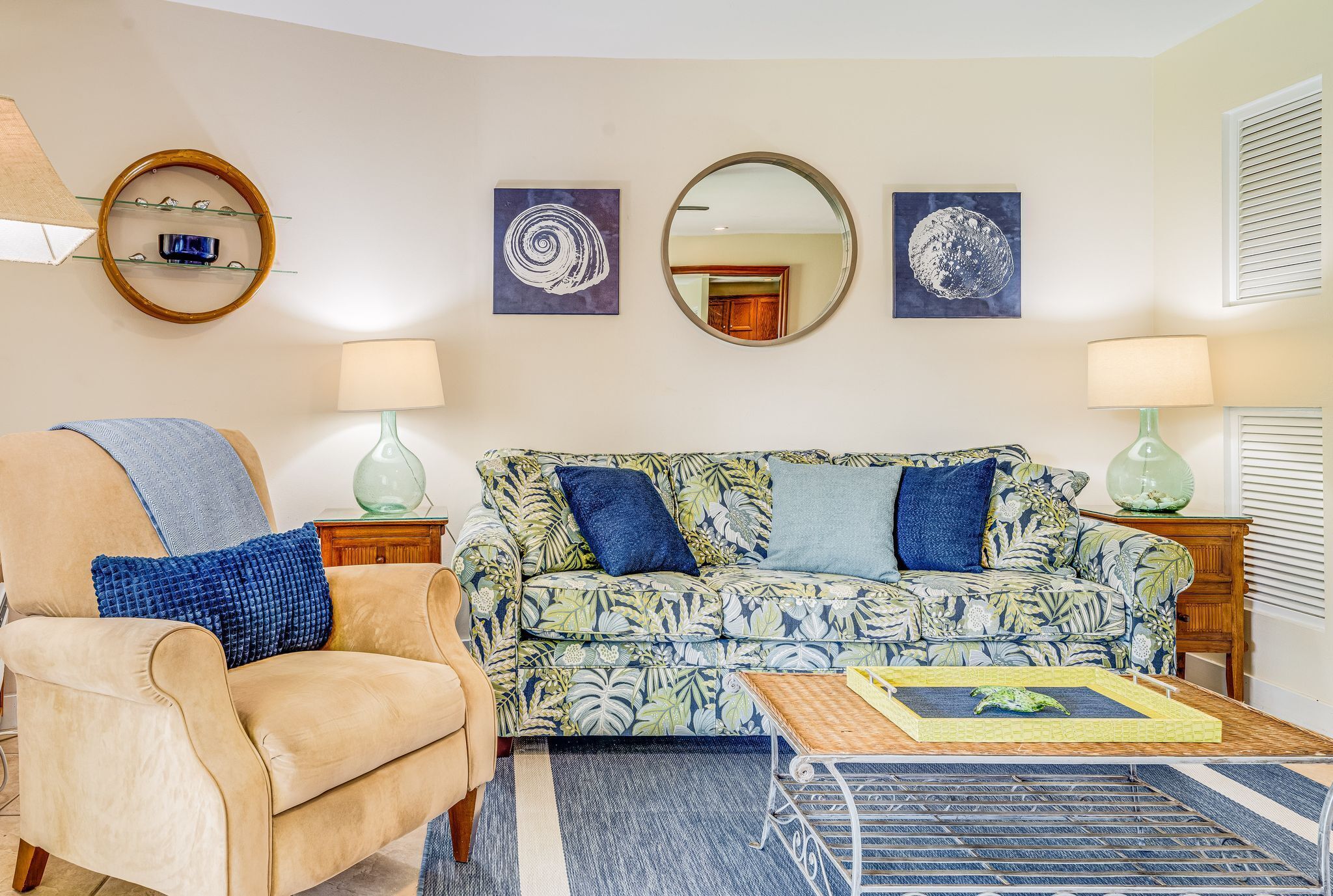 Ein Wohnzimmer mit einem beigefarbenen Sessel, einem sofa mit Blumenmuster und einem Couchtisch. Zwei Wandlampen und drei gerahmte Kunstwerke befinden sich über dem Sofa.