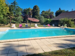 A swimming pool with lounge chairs and a covered area, surrounded by trees and a building.