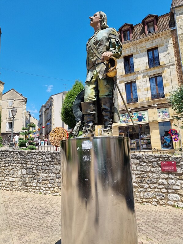 A statue of a historical figure in military attire, standing on a pedestal in an urban setting with buildings and shops in the background.