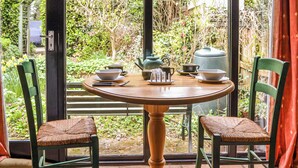 A round wooden table with two green chairs, a window with a view of a garden, and a door with a handle. - A lovely traditional Norfolk barn conversion in the heart of the coastline. (Sidestrand, near Cromer)