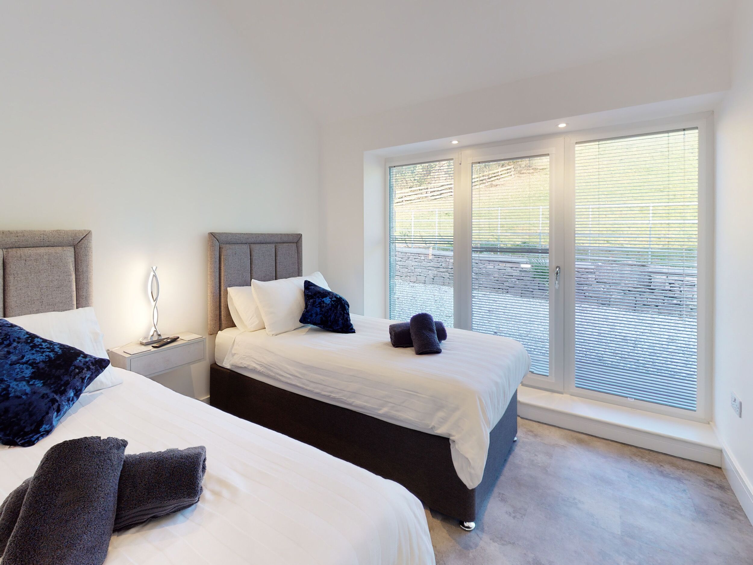 A double bed with a grey headboard, a bedside table with a lamp, and a large window with blinds.