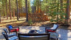 An outdoor seating area with wicker chairs and a glass-top table, surrounded by a forest and a stack of firewood. - Lovely Spring Home, Access to Pool (Black Butte Ranch)