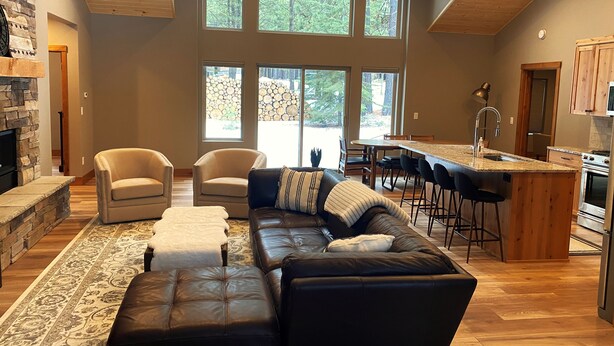 A spacious living room with a stone fireplace, leather sofas, and a large window overlooking a forest. - Lovely Spring Home, Access to Pool (Black Butte Ranch)