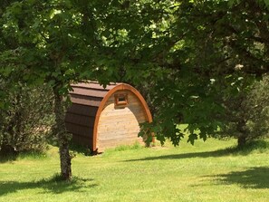 A wooden cabin with a sloped roof and a small window, surrounded by lush green trees and grass.