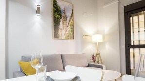 Ett modernt vardagsrum med en vit soffa, ett matbord med tallrikar och glas, en lampa och en inramad tavla på väggen.
