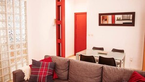 Een woonkamer met een grijze bank, een rode deur, een glazen tafel en een spiegel aan de muur.