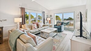 Ein geräumiges Wohnzimmer mit großem Fenster, Deckenventilator, einer Sofagarnitur, einem Couchtisch und einem Fernseher auf einem Schrank.