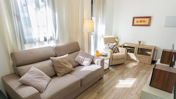 Ein Wohnzimmer mit einem beigen Sofa, einem kleinen Couchtisch und einer Lampe. Außerdem steht ein Sessel und eine Kommode mit Dekorationen dort.