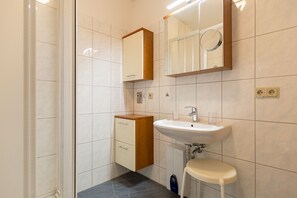 Un baño con lavamanos blanco, espejo, mueble de madera y un taburete pequeño.