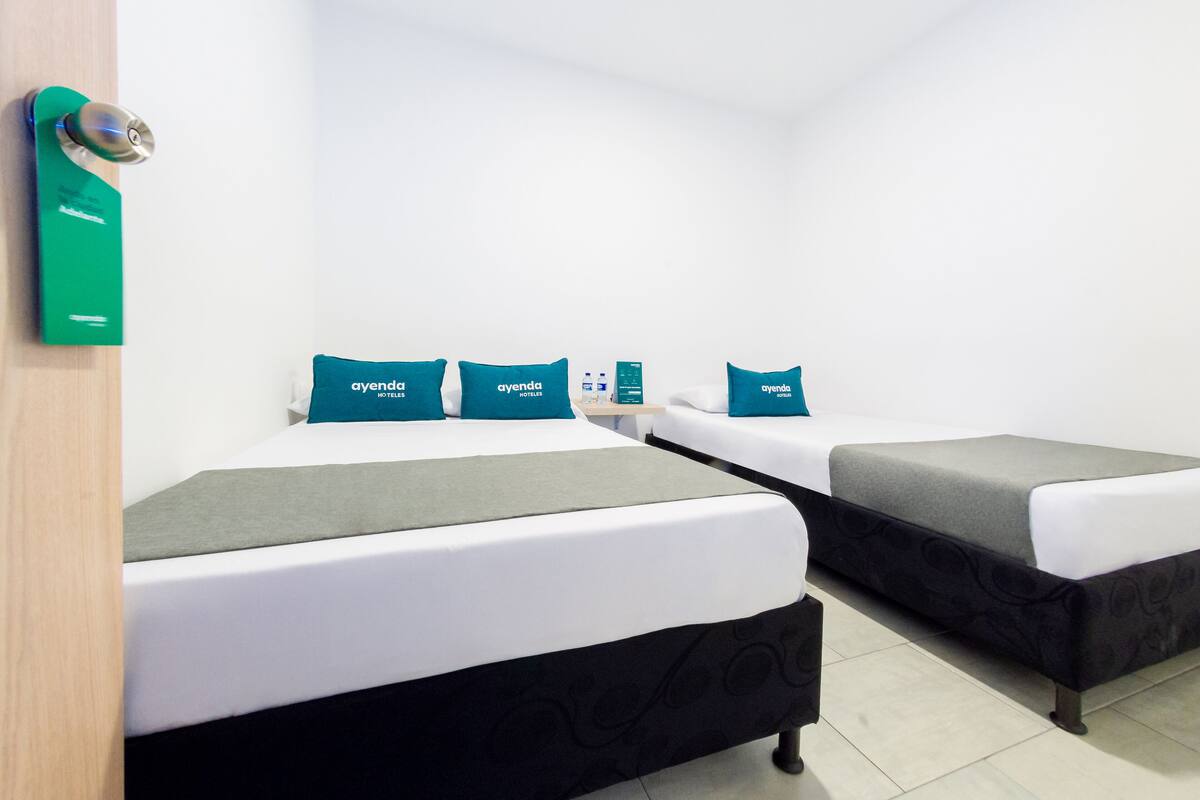 Une chambre d’hôtel avec deux lits, chacun agrémenté d’un oreiller turquoise et d’une couverture grise. On y trouve un mini-bar et une petite table placée entre les deux lits.